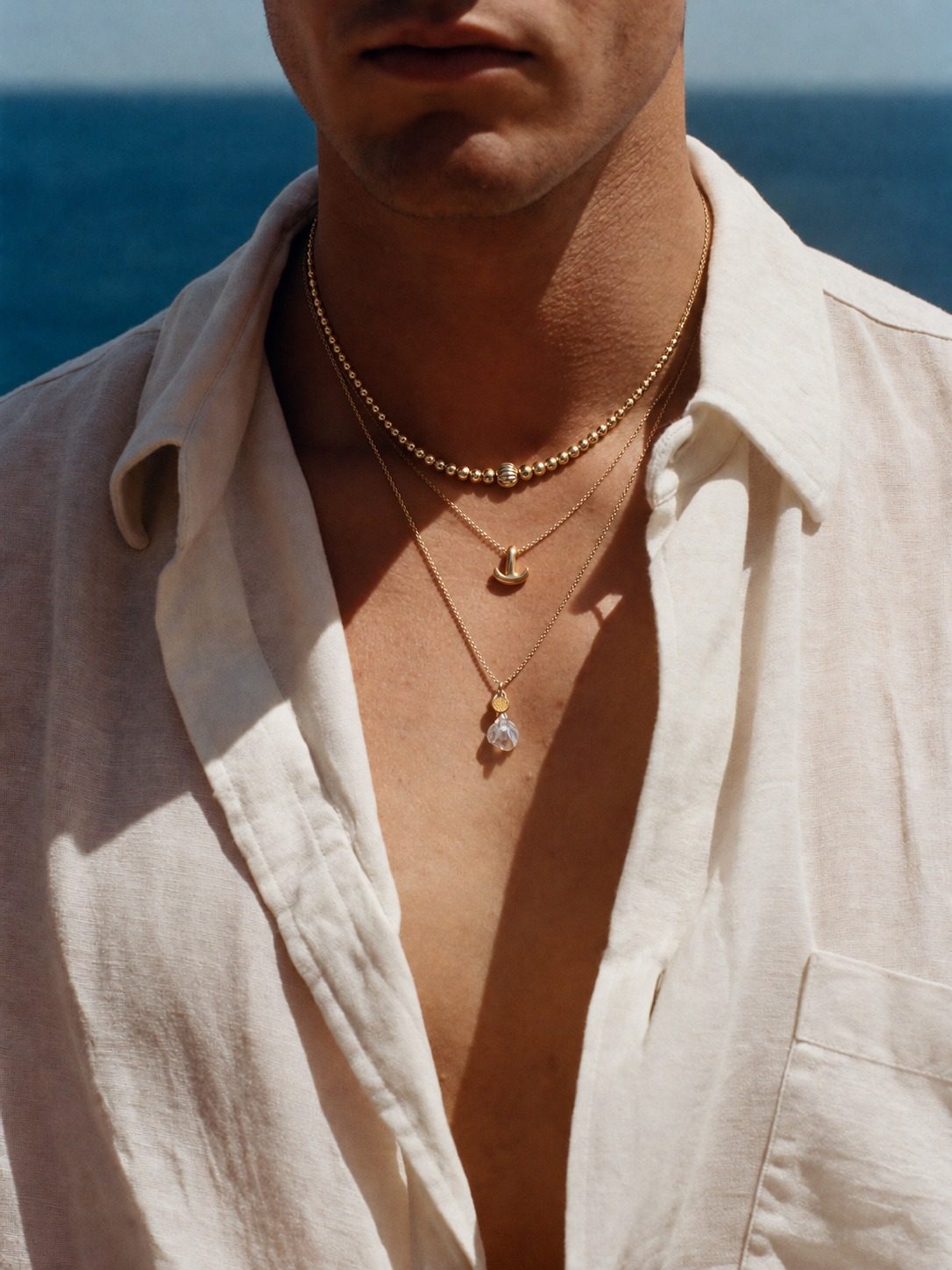 Cadenas de oro en capas con dijes, camisa de lino frente al mar